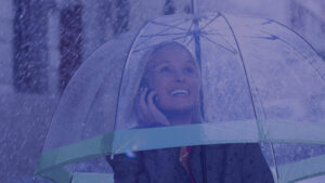 Smiling professional woman under a clear umbrella talking on her mobile phone during a rainstorm.